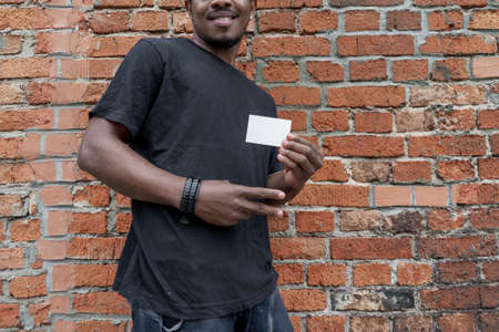 Dark-skinned man in T-shirt showing blank business card on bricked background.の写真素材