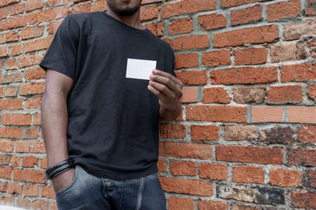 Dark-skinned man in T-shirt showing blank business card on bricked background.の写真素材