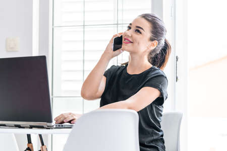 Young and pensive attractive woman working on laptop and talking by mobile phone in modern bright apartments. Freelance concept. Home office. Young entrepreneur.の写真素材
