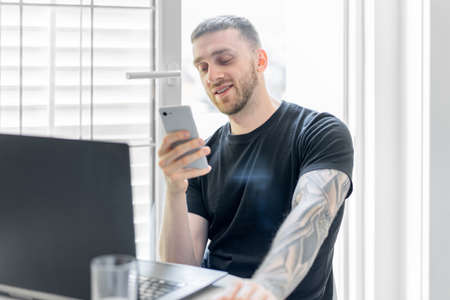 Young and pensive attractive man working on laptop and chatting by mobile phone in modern bright...