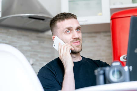 Young and pensive attractive man working on laptop and talking by mobile phone in modern bright apartments. Freelance concept. Home office. Young entrepreneur.の写真素材
