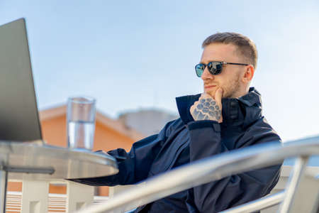 Handsome pensive stylish man sitting on balcony and working on laptop. Home office. Freelance concept. Young entrepreneur.の写真素材