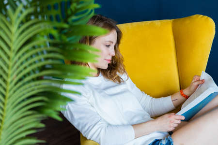 Young blonde woman reading book at home and sitting in yellow armchairの写真素材