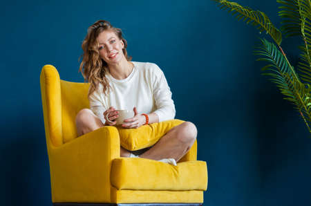 Blonde woman reading book at home, drinking coffee or tea and sitting in armchairの写真素材