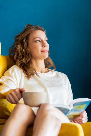 Blonde woman reading book at home, drinking coffee or tea and sitting in armchairの写真素材