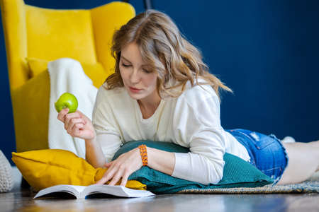 Blonde woman reading book at home and laying on the floor next to yellow armchairの写真素材