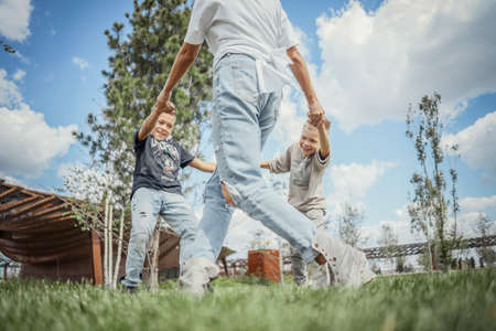 Young mum turning around, whirling and having fun with her sons at green modern park. Happy family concept.の写真素材