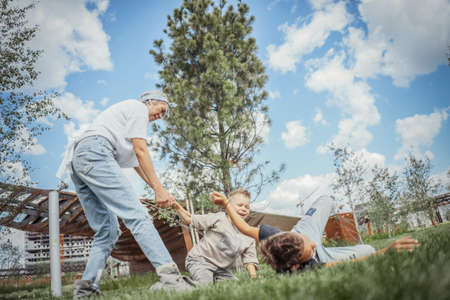 Young blonde mum turning around, whirling with her sons at park. Happy family concept. Family is on foreground and focus, park is blurred.の写真素材