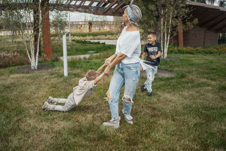 Young mum turning around, whirling and having fun with her sons at green modern park. Happy family concept.の写真素材