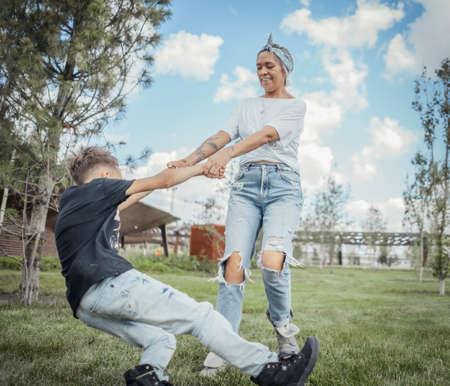 Young pretty mum turning around, whirling with her son at park. Happy family concept. Family is on foreground and focus, park is blurred.の写真素材