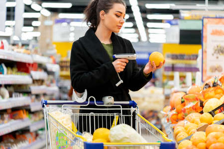 Young attractive woman holding bar code scanner and scanning products in store. Concept of modern gadgets and devices, Fruits and vegetables department. Healthy lifestyle. loosing weight.の写真素材