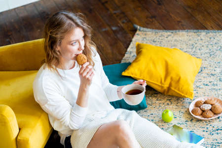 Young blonde woman reading book at home and laying on the floor with cushions next to yellow armchair. Girl eating cookie and green apple. Cozy and comfortable timespending.の写真素材
