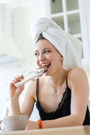 Woman eating chocolate in white bright apartments. Diet keeping concept. Loosing weight. Woman having breakfast after taking a shower with towel on her head. Cheat meal.の写真素材