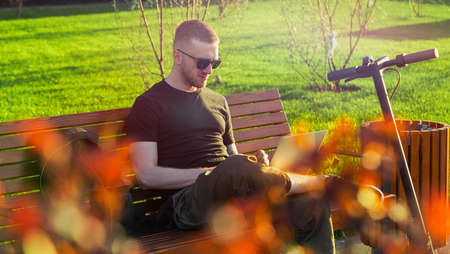 Young attractive man with laptop and modern electric kick scooter sitting at contemporary park at bench. and working Freelancer or business man working on laptop. Beautiful Sunset time.の写真素材