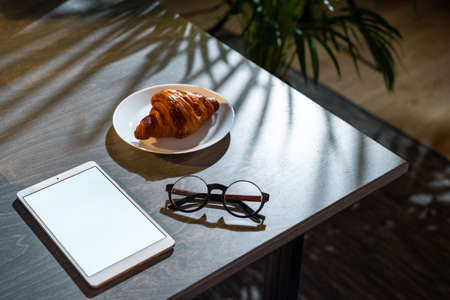 Close up of croissant, glasses and pad with blank screen on dark wooden table with tropic plant on background.の写真素材
