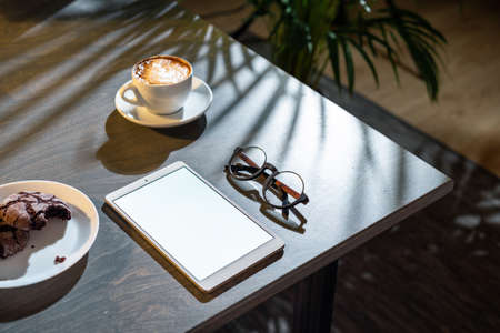 Close up of white coffee cup, oat cookies, glasses and pad with blank screen on dark wooden table with tropic plant on background.の写真素材