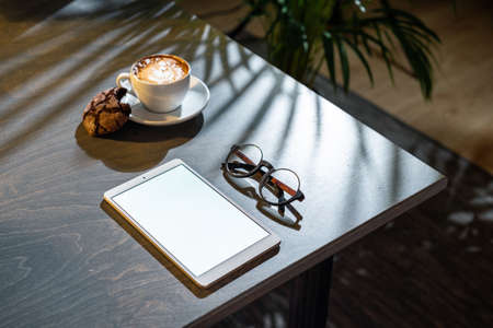 Close up of white coffee cup, oat cookies, glasses and pad with blank screen on dark wooden table with tropic plant on background.の写真素材