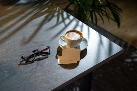 Close up of white coffee cup, glasses and business cards on dark wooden table with tropic plant on background.の写真素材