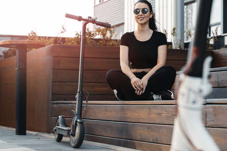 Young attractive woman sitting next to electrick kick scooter at modern park.の写真素材