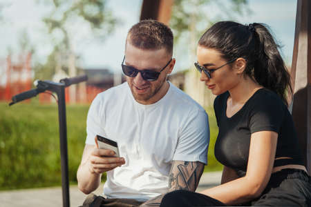 Young man and woman sitting in park and looking at mobile phone screen.の写真素材