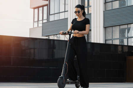 Young attractive woman riding electrick kick scooter at modern cityscape.