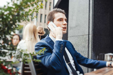 Young pensive attractive businessman talking by mobile phone in street cafe and looking straight.の写真素材