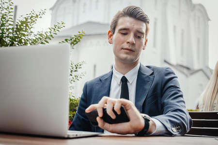Close up of businessman sitting next to laptop, holding mobile phone in hand and looking at watches.の写真素材