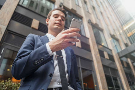 Close up of businessman next to skyscraper at downtown holding mobile phone and looking at screen. Bottom viewの写真素材