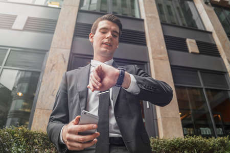 Close up of businessman next to business center at downtown holding mobile phone in hand and looking at watches. Bottom viewの写真素材
