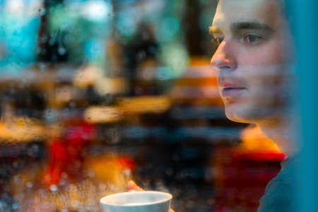Young pensive handsome man drinking tea or coffee on rainy day and looking through the window.の写真素材