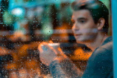 Young pensive handsome man drinking tea or coffee on rainy day and looking through the window.の写真素材