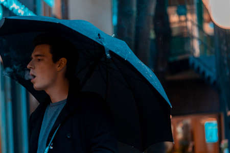 Young pensive handsome man with umbrella on rainy day going for a walk in park.