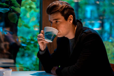 Young pensive handsome man drinking tea or coffee on rainy day and looking through the window.の写真素材