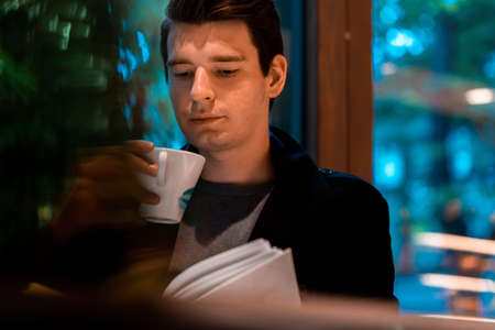 Young pensive handsome man drinking tea or coffee on rainy day and looking through the window.の写真素材