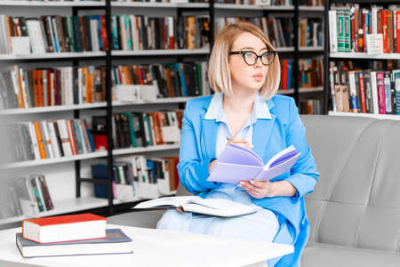 Young pensive attractive woman in formal attire makes some notes in notepad at library.の写真素材