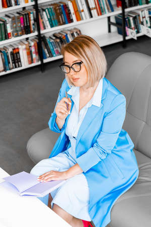 Young pensive attractive woman in formal attire makes some notes in notepad at library.の写真素材