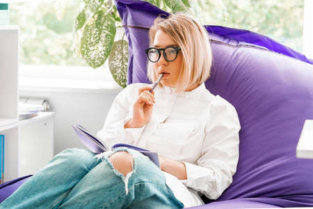Young attractive smiling woman makes some notes in notepad at library and sitting on violet couch.の写真素材