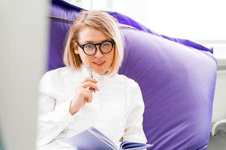 Young attractive smiling woman makes some notes in notepad at library and sitting on violet couch.の写真素材