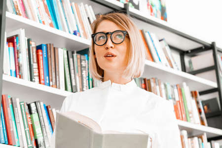 Young pensive attractive woman with glasses in formal clothes reading a book at library.の写真素材