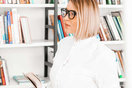 Young pensive attractive woman with glasses in formal clothes reading a book at library.の写真素材