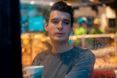 Young pensive handsome man drinking tea or coffee on rainy day and looking through the window.の写真素材