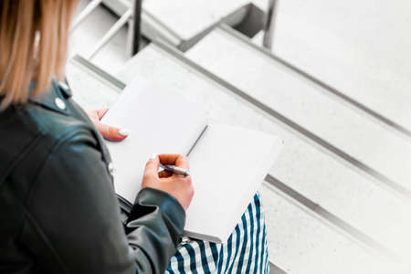 Young attractive blond woman makes some notes in violet notepad at library and sitting on ladder steps.の写真素材