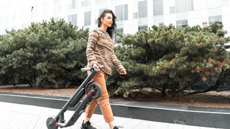 Young attractive smiling woman holding electric kick scooter while going for a walk at street. light weight and transportable compacted scooter. Urban cityscapeの写真素材