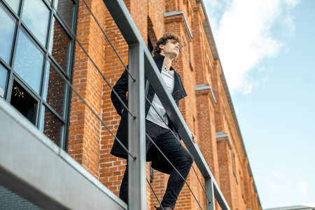 Young attractive pensive man standing on staircase next to banister on red bricked building background.の写真素材