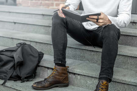 Close up of pensive man sitting on concrete steps of red bricked buildingand holding notepad in hands.の写真素材