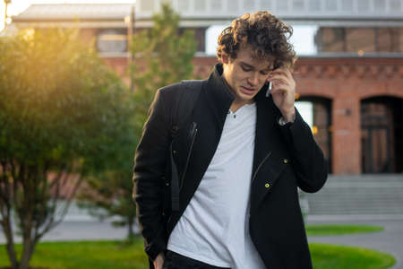 Close up of man in black coat talking by mobile phone while going through red bricked modern buildings and green trees. Cityscape urban background.の写真素材