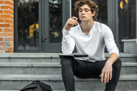 Attractive pensive student sitting on modern building steps and making notes in his black notepad.の写真素材