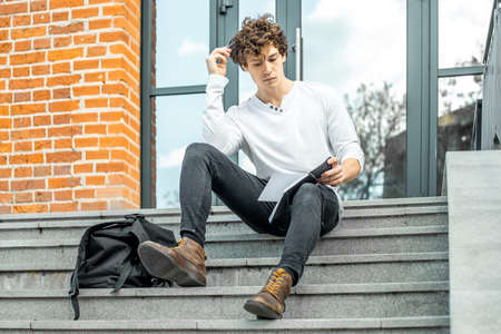 Attractive pensive student sitting on modern building steps and making notes in his black notepad.の写真素材