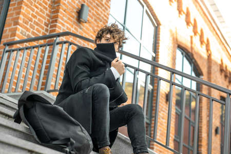 Young attractive pensive man sitting on concrete steps of modern red bricked building and wrapping up in black coat.の写真素材