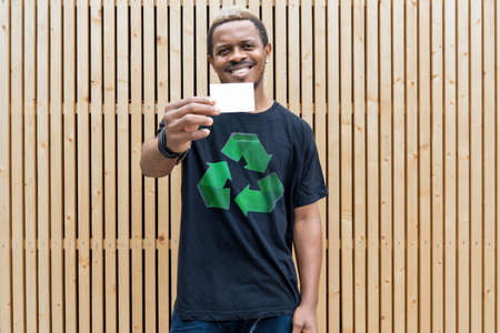 Young attractive man in t-shirt with recycle symbol on wooden background holding blank business card. Eco symbol.の写真素材